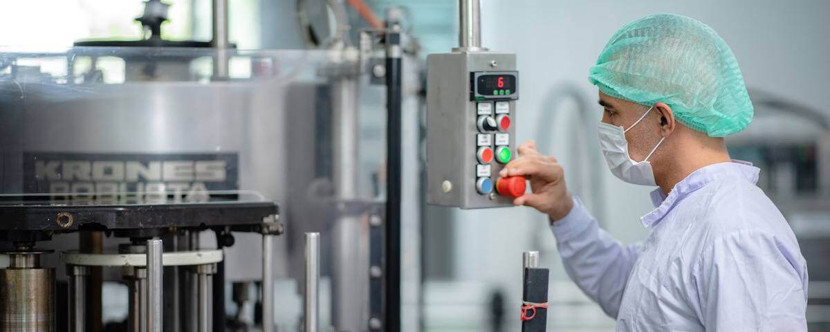 A technician wearing a hairnet and face mask operates a control panel in a clean manufacturing environment