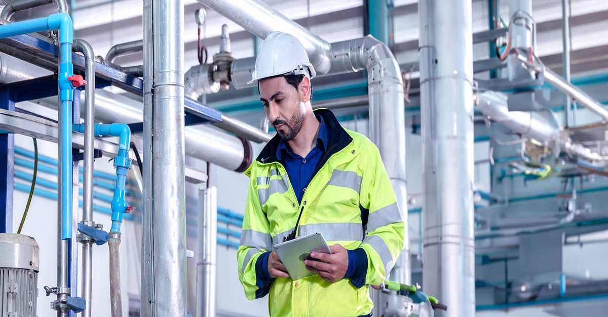 An engineer inspecting industrial piping and taking notes on a tablet
