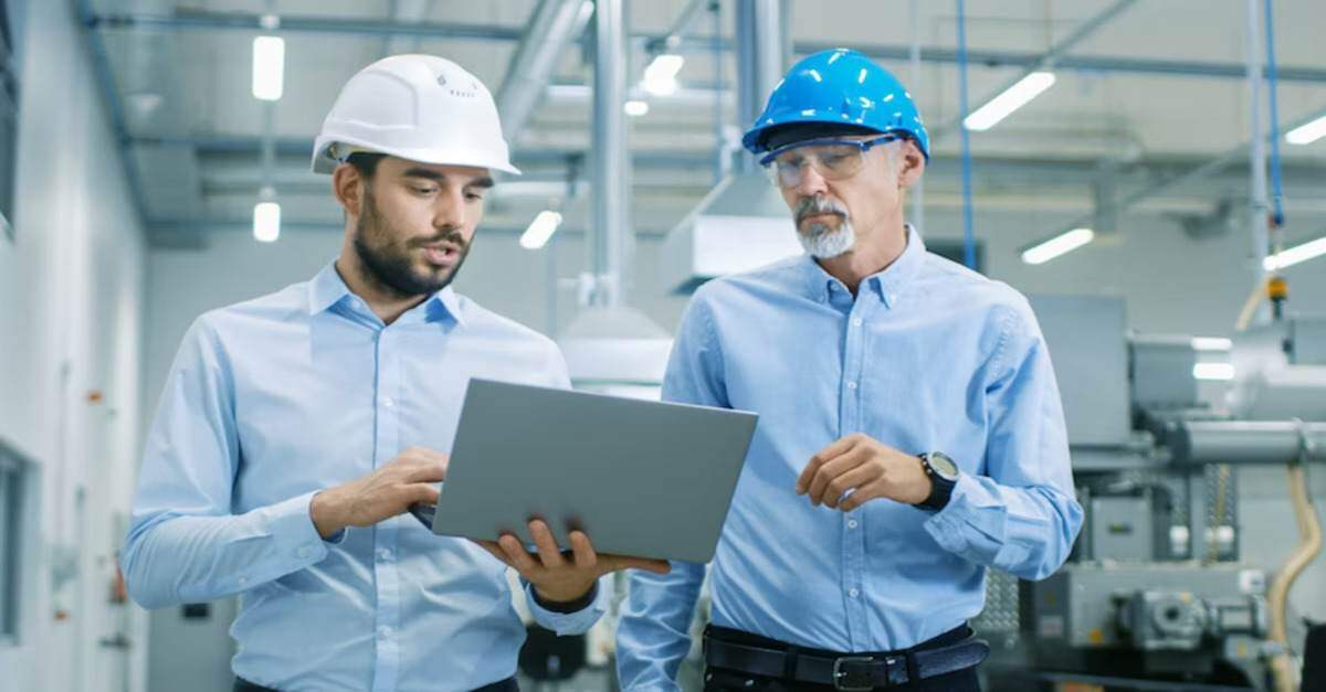Two engineers in hard hats discussing work while looking at a laptop in an industrial facility