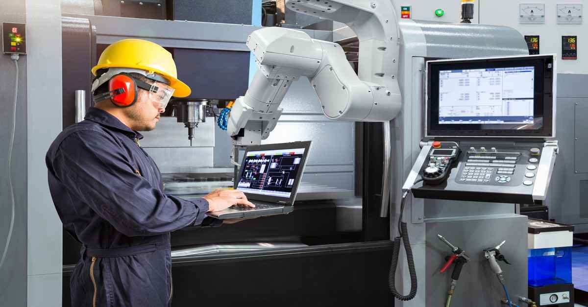 Engineer in safety gear operating a laptop while monitoring robotic machinery in an industrial setting