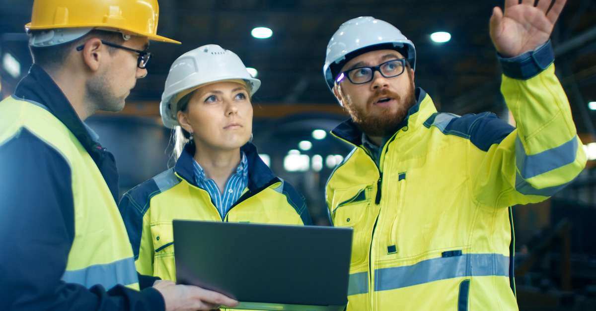 Three engineers in high-visibility jackets and hard hats discussing a project in an industrial facility
