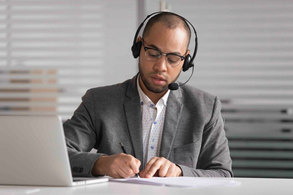 Man in a suit wearing a headset, taking notes while working on a laptop