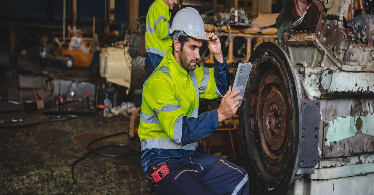 Industrial worker wearing safety gear, inspecting machinery with a tablet