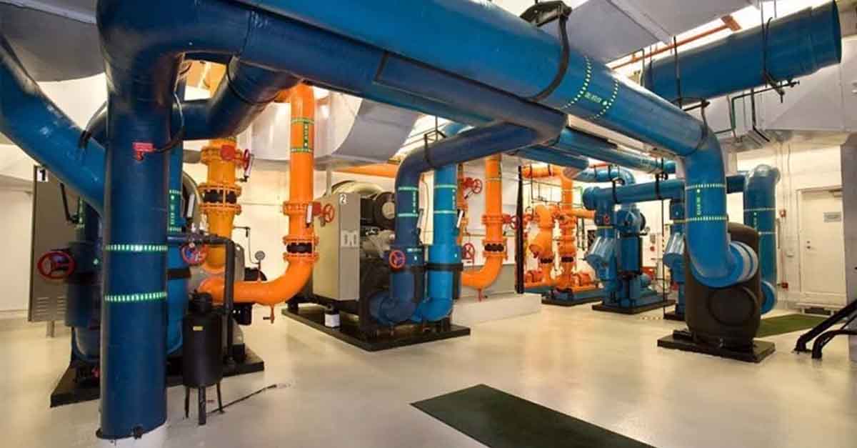 A brightly lit industrial facility with large blue and orange interconnected pipes, valves, and machinery, indicating a complex water or HVAC system.