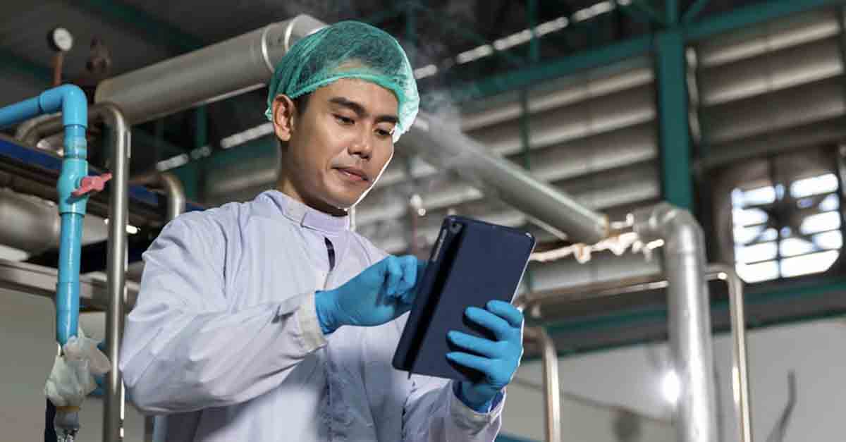 A man in a lab coat is using a tablet in an industrial setting
