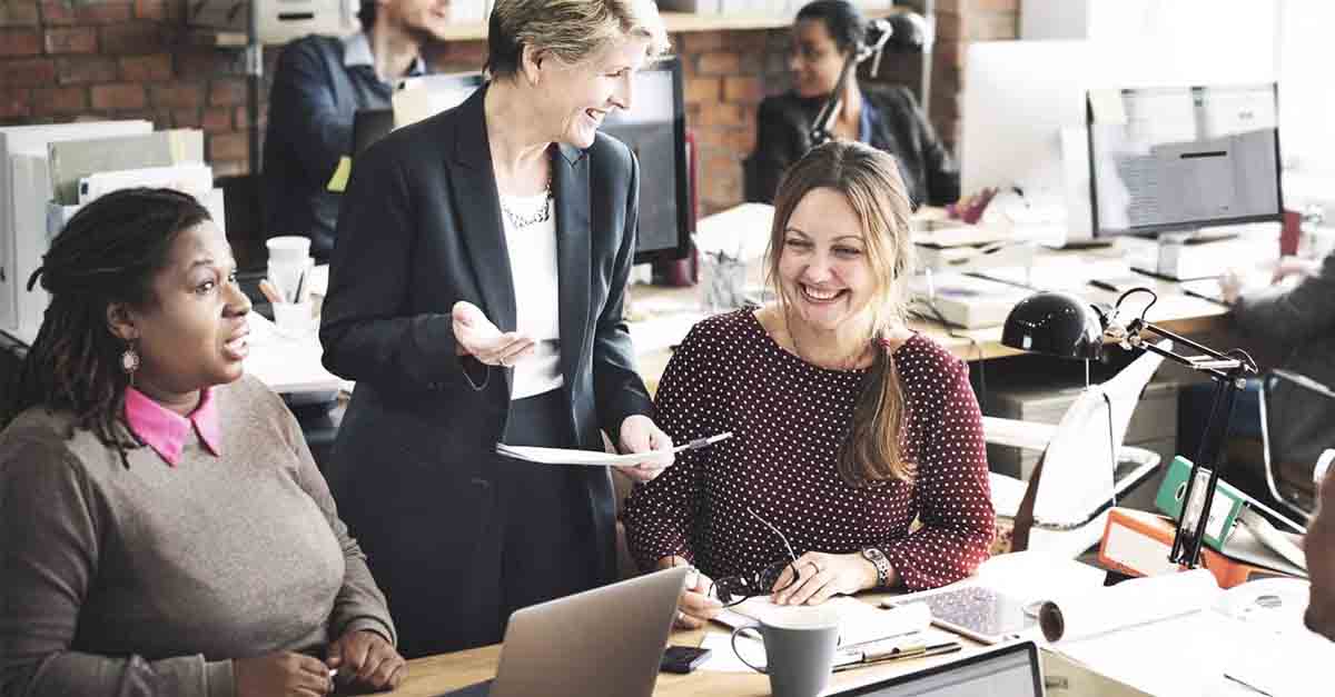 Group of office workers smiling and talking around a desk in a busy workspace
