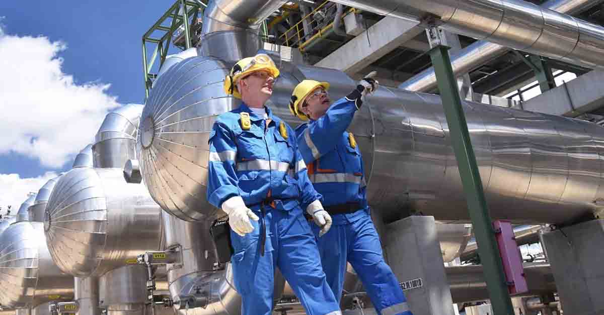 Two workers in blue uniforms inspecting an industrial plant with large metal pipes