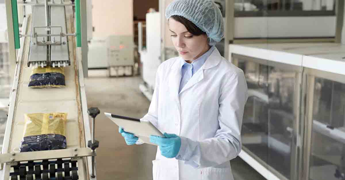 Worker in a lab coat using a tablet to inspect a conveyor belt in a packaging facility