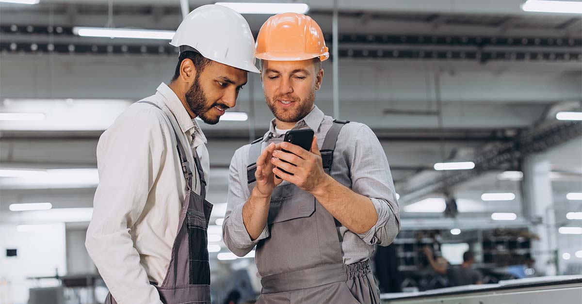 Two factory workers wearing hard hats looking at a smartphone inside an industrial facility