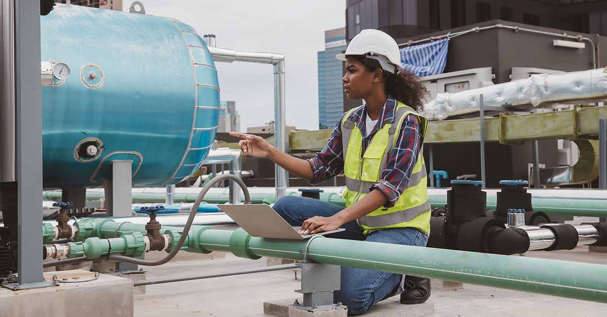 Female technician inspecting equipment outdoors while using a laptop and pointing at a pipe