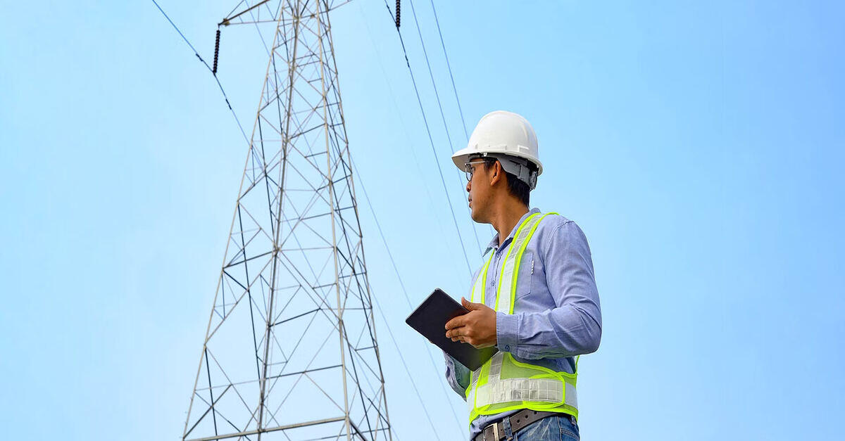 A construction worker holding a tablet while standing near a tall power transmission tower