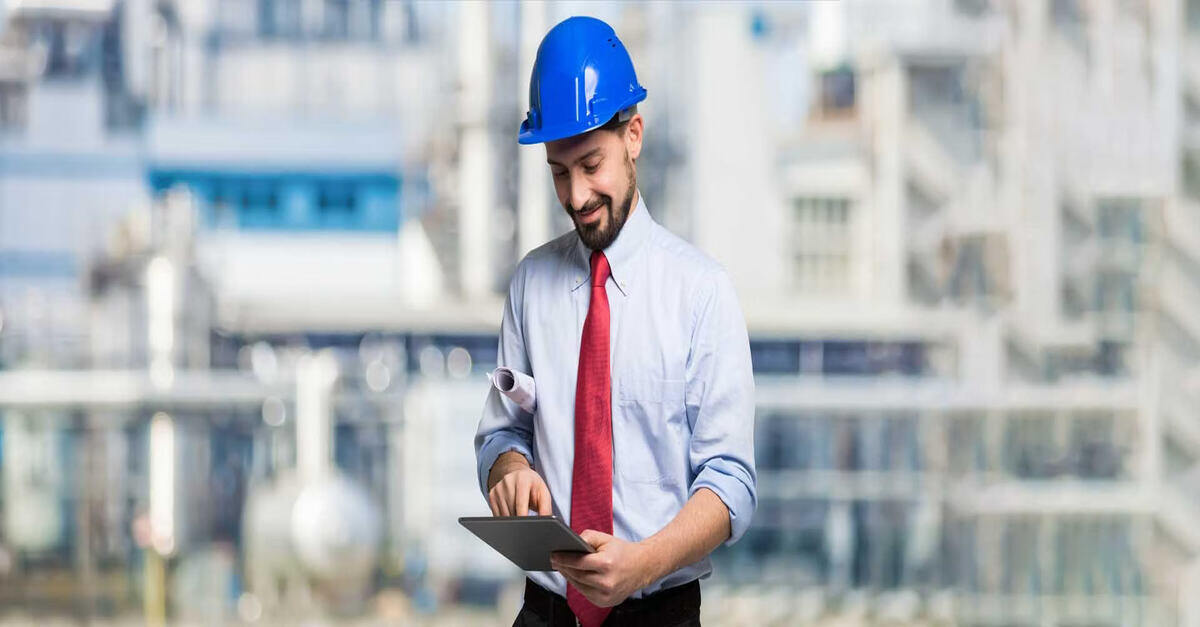 An engineer wearing a blue hard hat and a red tie stands in front of an industrial facility, smiling as he works on a tablet