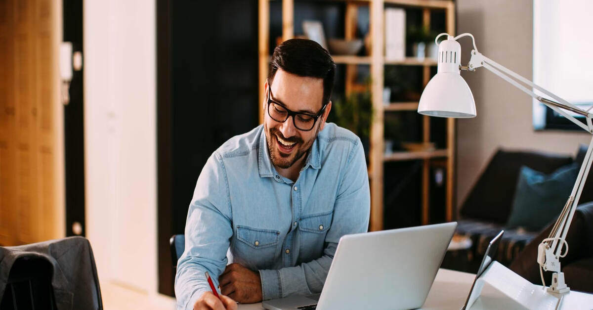 Man working at a desk with a laptop, smiling while writing in a notebook