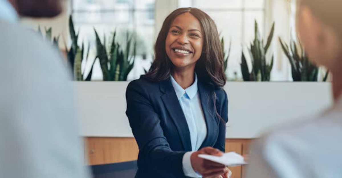 A smiling woman in a business suit hands a document to someone during a professional meeting